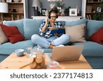 Small photo of Young beautiful hipster woman sitting on sofa at home listening music on wireless headset and reading a book. Freelance female enjoy free time after finishing work on laptop, read new novel bestseller