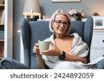 Simple living. Senior woman sitting alone on chair at home drinking tea or coffee enjoying her time and life. Older female thinking about her past and happiness, satisfied with her life decisions