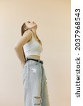 Small photo of Low-angle image of attractive woman in baggy ripped jeans with belt standing half-turned to camera looking up with head titled back and hands crossed behind her back, feeling nervous and anxious