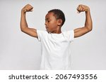 Small photo of Indoor studio shot of strong elementary school kid with dark skin standing in front of camera showing his biceps and tense muscles, putting hands bent in elbows up, feeling proud of himself