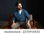 Happy joyful married guy with breaded chin having rest sitting on brown small couch, looking aside with toothy smile, putting hands on knees, wearing blue classical cardigan and stylish pants