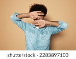 Man introvert hiding face behind opened palms, feeling scared or ashamed of his mistakes and behavior, dressed in denim clothes isolated on brown studio background. Embarrassment and shyness