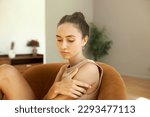 Side view portrait of sad melancholic beautiful caucasian girl coping with grief, looking down touching her shoulder sitting on couch in her living-room. Negative human emotions and feelings