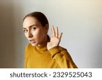 Portrait of concentrated anxious female eavesdropping holding head next to ear, listening carefully to strange sounds, feeling scared, standing against gray studio background with opened mouth