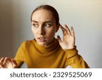 Portrait of anxious scared female stepping carefully eavesdropping with anxious facial expression, listening to strange scary sounds attentively, holding hand around ear isolated on gray background