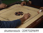 Small photo of a carrom game in progress. Players’ hands flick the striker on a traditional Indian board. The wooden board with white and black pieces is the focus The game embodies culture, leisure, and strategy.