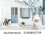 Exterior of a white mediterranean-style house with a blue door and a window, a flowering tree. Traditional patio of Santorini