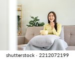 Young asian woman putting a bowl of popcorn on pillow and eating popcorn while sitting on the big comfortable sofa to watching movie on television in living room at home.