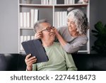 Senior woman putting glasses on beloved while sitting on the couch to watching movie on tablet and spending time to doing activity for relaxation in life retirement together at living room of home.