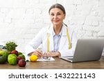 Indoor shot of positive smiling Caucasian female nutritionist in her sixties working online, sitting in front of open laptop pc, consulting clients on nutrition via social networks, making notes
