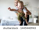 Pretty girl babysitting adorable toddler, having fun in stylish interior. Portrait of beautiful happy young Caucasian mom enjoying precious time with her cute baby son, holding him like flying plane
