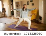 Full length shot of happy energetic mature woman in casual clothes exercising at home because of social distancing, practicing yoga on mat, standing in warrior ii pose. Age, wellness and health