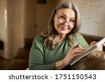 Stylish creative middle aged female writer or blogger in glasses sitting comfortably on couch with copybook and pen, handwriting, making notes, putting down ideas, thoughts, smiling at camera