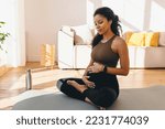 Pregnant african woman in sports clothes sitting on mat in her living-room, having rest after doing prenatal fitness training exercise, stroking her belly, feeling pushes of her baby