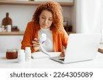 Small photo of Redhead nutritionist working from home on laptop at her article about deficiency of food supplements and its influence on human body and health sitting at kitchen table with vitamin bottle