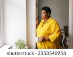Side view of sad african american overweight female in yellow clothes and glasses standing next to window with cup of hot drink, grandma missing her grandchildren, waiting them to visit her