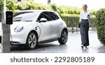 Small photo of Businesswoman using tablet, walking while recharging her electric vehicle with charging station at public car parking. Progressive lifestyle of technology and ecological concern by EV car