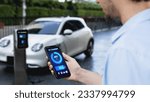Small photo of Businessman holding smartphone display battery status interface by smart EV mobile application while EV car recharging electricity from charging station in car park. Peruse