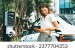 Small photo of Young woman travel with EV electric car charging in green sustainable city outdoor garden in summer. Urban sustainability lifestyle by green clean rechargeable energy of electric BEV vehicle innards