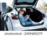 Small photo of Little boy sitting on car trunk, using smartphone while recharging eco-friendly car from EV charging station. EV car road trip travel as alternative vehicle using sustainable energy concept. Perpetual