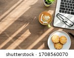 Wood office desk table with dish of cookies, cup of latte coffee and supplies. Top view with copy space, flat lay. Working from home in the morning concept.