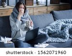 Ill brunette girl is sitting at sofa and working remotely on laptop. Female is blowing out snot, having fever and headache. Young woman is treated at home, took sick leave. Winter cold and flu concept