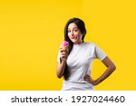 Pretty Indian girl or young Asian woman eating strawberry ice cream in cone against yellow studio background