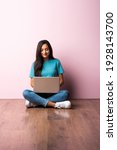 Small photo of Indian asian young woman or girl sitting with laptop on her lap against pink wall on wooden floor