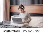 Young asian woman in casual clothes with headphone put laptop on pillow to looking on laptop screen and working while relaxing by listening to music and sitting on the bed in home bedroom