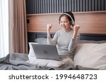 Young asian woman with headphone put laptop on pillow to working about business and raising hand when finish work while relaxing by listening to music and sitting on the bed in home bedroom