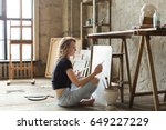Woman painter sitting on the floor in front of the canvas and drawing. Artist studio interior. Drawing supplies, oil paints, artist brushes, canvas, frame. Workshop or art class. Creative concept