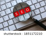 Selective focus of magnifying glass and computer keyboard with red key written with SEO Search Engine Optimization on a wooden background.