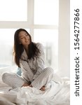 Small photo of Young beautiful Asian woman woke up in sunny morning. Attractive girl in nightgown sitting on bed at home against big window. Gorgeous model with closed eyes