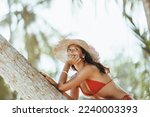 Summer lifestyle funny portrait of cute young bikini girl with tanned perfect body in red swimsuit, posing sitting on palm tree and looking away at the tropical beach resort during sea vacations. 