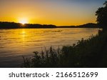 Tranquil sunset on Missouri River; trees and plants in foreground; forest in background; setting sun reflects in water