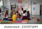 Small photo of children sitting on floor while caring teacher explains lesson using toy in kindergarten. Elementary school students and teacher sit in circle in classroom