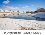 Snow around a lake in winter. Houses line the shore. Footprints can be seen in the snow. The sky is bright blue with puffy clouds. The water is a dark blue green.