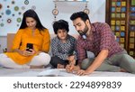 Small photo of A handsome educated father helping his adorable son in completing his homework - Indian nuclear family in their bedroom . A smiling Indian mother browsing her mobile while sitting on the bed - elec...