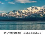 Small photo of Disenchantment Bay, Alaska, USA - July 21, 2011: Yakuta bay, sun lights up snow on mountains along ocean shore under blue cloudscape. Blue ocean water up front. Forested shoreline on horizon