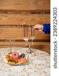 Small photo of A woman's hand with a shamash candle lights the Hanukkah candles at the table with donuts. Vertical photo.