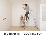 Young adult couple making interior change. Man standing on metal ladder and woman help applying new wallpaper on white wall in room. Working together. Repair work of home. 