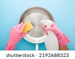 Woman hands in pink rubber protective gloves holding white detergent bottle, yellow sponge and washing stainless frying pan on light blue table background. Closeup. Pastel color. Point of view shot.