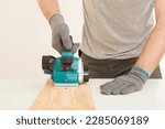 Small photo of Young adult man hands using electric jointer and shaving old wooden plank for furniture or floor on white table at light gray wall. Closeup. Preparing material for repair work of home. Front view.