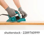 Small photo of Adult man hands using electric jointer and shaving old wooden plank for furniture or floor on white table at light gray wall background. Closeup. Preparing material for repair work of home. Side view.