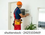 Small photo of Electrician with screwdriver tighten up switching electric actuator equipment in fuse box. View from the back, a young specialist in a yellow hard hat and overalls. Indoor