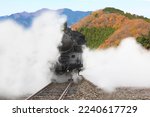 A steam locomotive emitting white smoke with a backdrop of mountains filled with autumn leaves