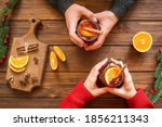 Christmas hot drink - mulled red wine in glass mug with spices and fruits in the hands of men and women. New Year decoration on a wooden background. Top view
