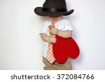 Little boy in hat and tie holding a red paper heart, lowering his gaze and looking confusedly, isolated portrait on white background, romantic and love concept