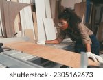 Small photo of Female carpenter using saw machine for cutting sawn plywood during working in wood workshop. Female joiner wearing safety uniform and working in furniture workshop