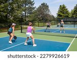 A competitive doubles game of pickleball at the net on a blue and green court in summer.